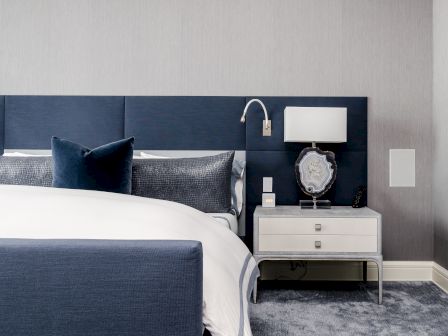 A modern bedroom with a navy and gray color scheme, featuring a bed, a stylish nightstand with a lamp, and a decorative wall piece.