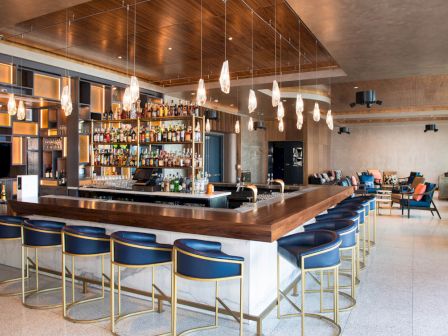 The image shows a modern bar with blue bar stools, pendant lights, and a fully stocked liquor shelf. There's seating in the background area.