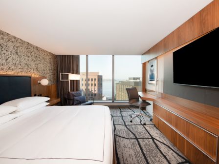 A modern hotel room with a large bed, a desk, a flat-screen TV, and a window offering a cityscape view completes the scene.