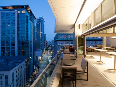 A modern rooftop terrace overlooks a city street with tall buildings at dusk. There are tables and chairs along the edge.