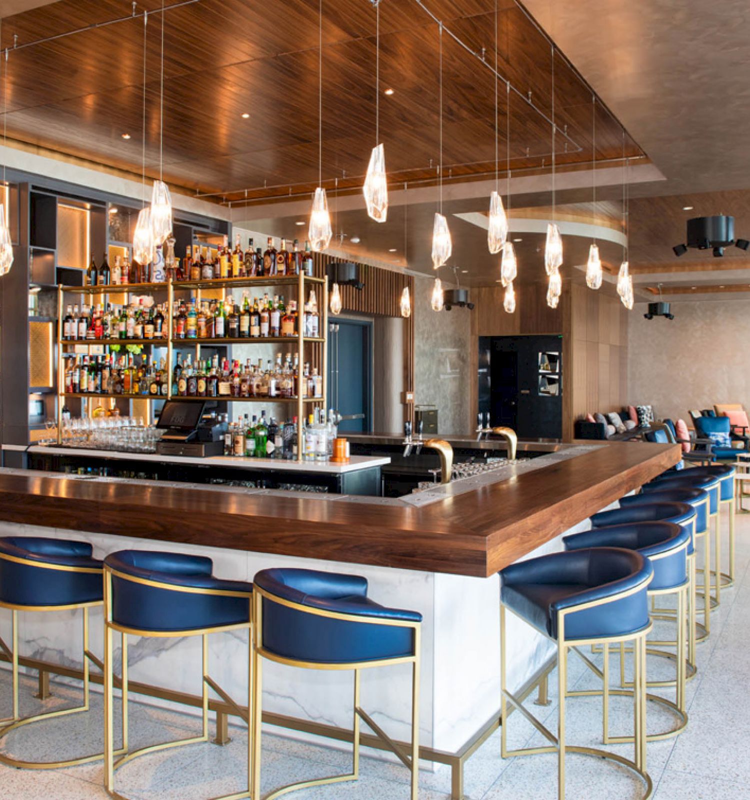 A modern bar with a wooden counter, blue stools, hanging lights, and a well-stocked liquor shelf in a spacious, elegantly designed room.