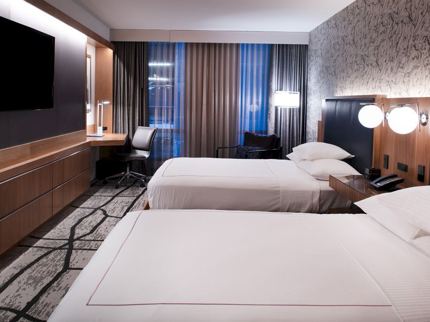 A modern hotel room with two beds, a wall-mounted TV, desk, chair, and stylish decor, featuring a patterned carpet and curtains.