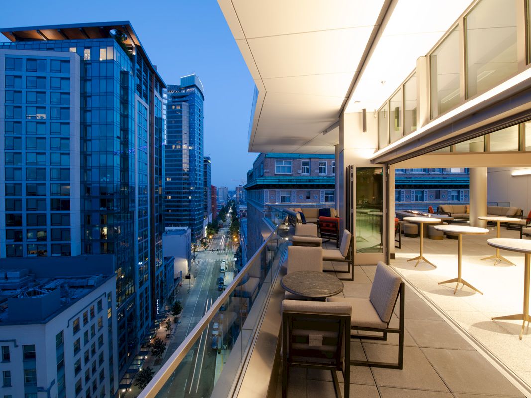 A modern rooftop terrace overlooking a cityscape with tall buildings and evening lights, featuring seating and tables for relaxation.