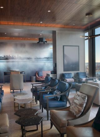 The image shows a modern lounge with plush seating, large windows offering a scenic view of the water, and a mural of a cityscape on the wall.