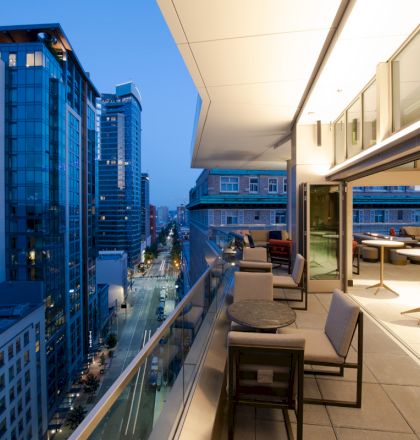 A modern rooftop balcony overlooking a cityscape at dusk, featuring outdoor seating and warm interior lighting.