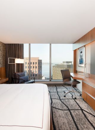 The image shows a modern hotel room with a large bed, desk, chair, TV, and window with a city view, featuring wooden accents throughout.