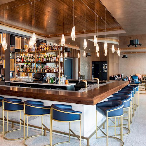The image shows a modern bar with blue bar stools, a stocked back shelf, hanging lights, and seating area in the background.