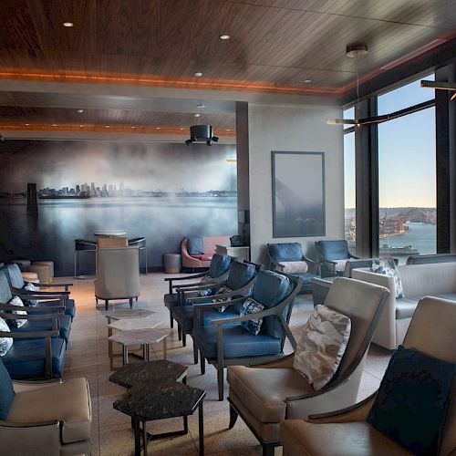 A modern lounge with comfortable seating, large windows showing a waterfront view, and a wall mural of a city skyline enhances the decor.