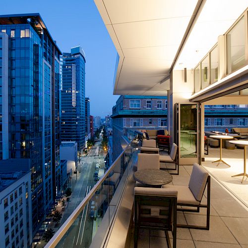 A modern cityscape is visible from a stylish rooftop patio with tables and chairs, overlooking a street flanked by tall buildings at dusk.