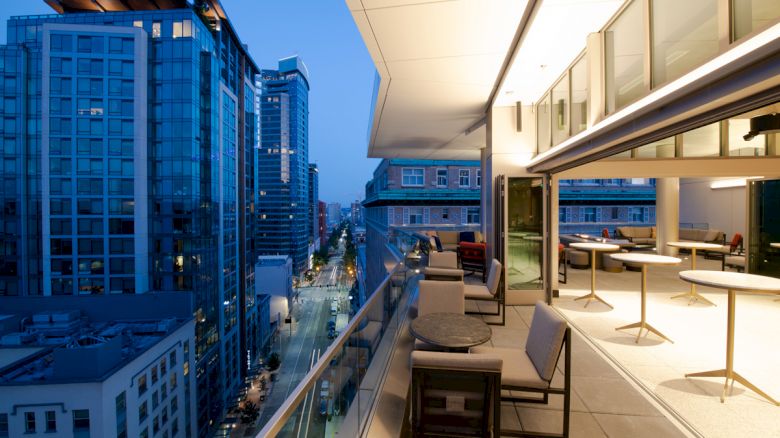 A modern urban balcony with tables and chairs overlooks a cityscape at dusk, featuring tall buildings and a street below.