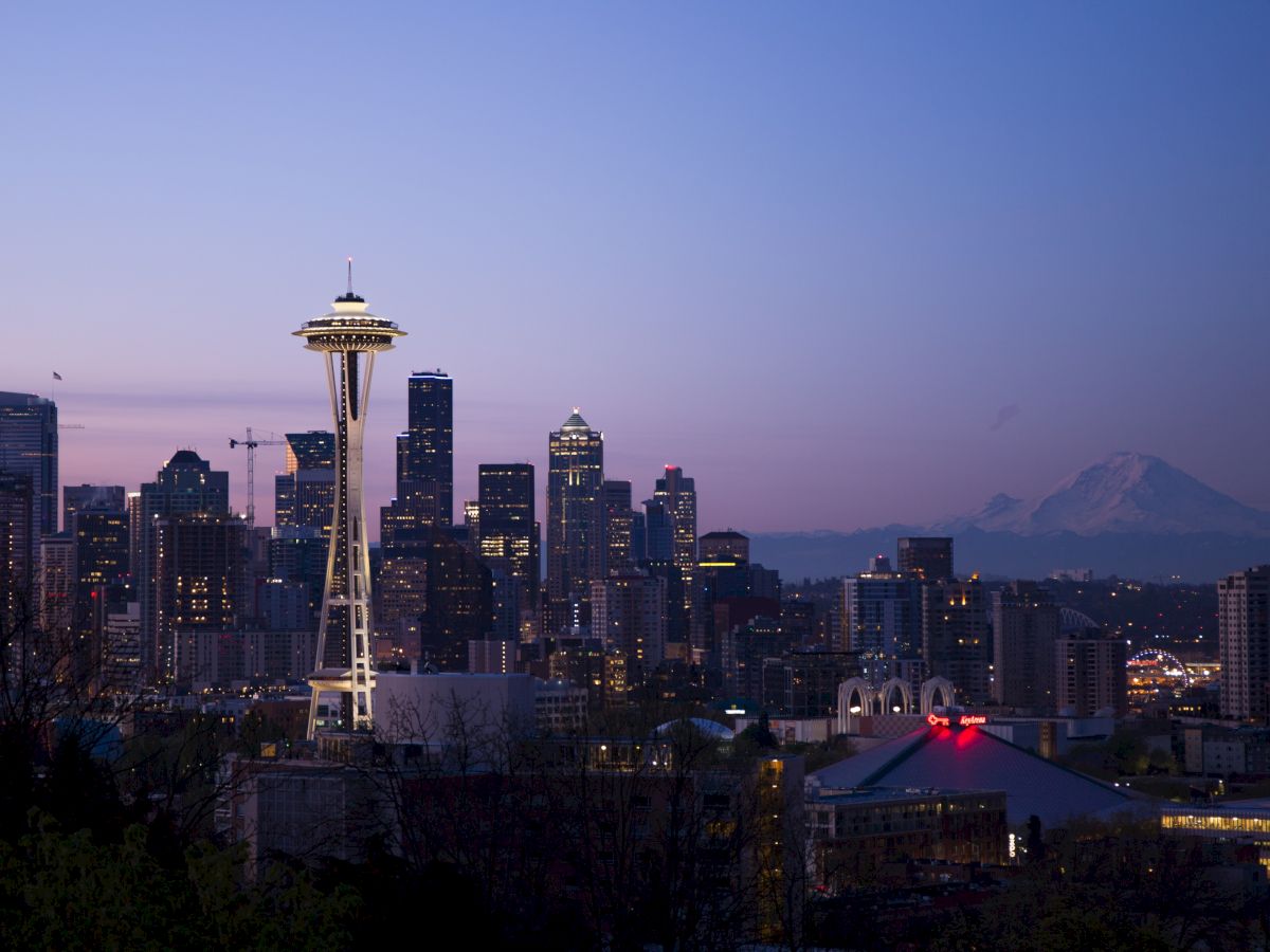 Skyline of Seattle at dusk, featuring the Space Needle and city buildings, with Mount Rainier in the background under a purple sky.
