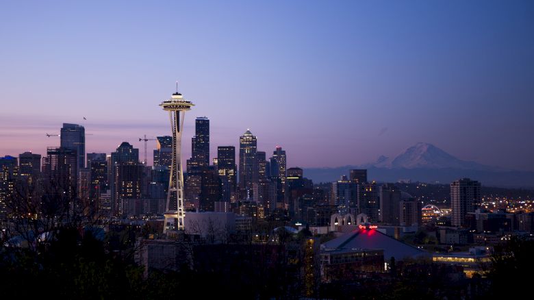 Skyline of Seattle at dusk, featuring the Space Needle and city buildings, with Mount Rainier in the background under a purple sky.