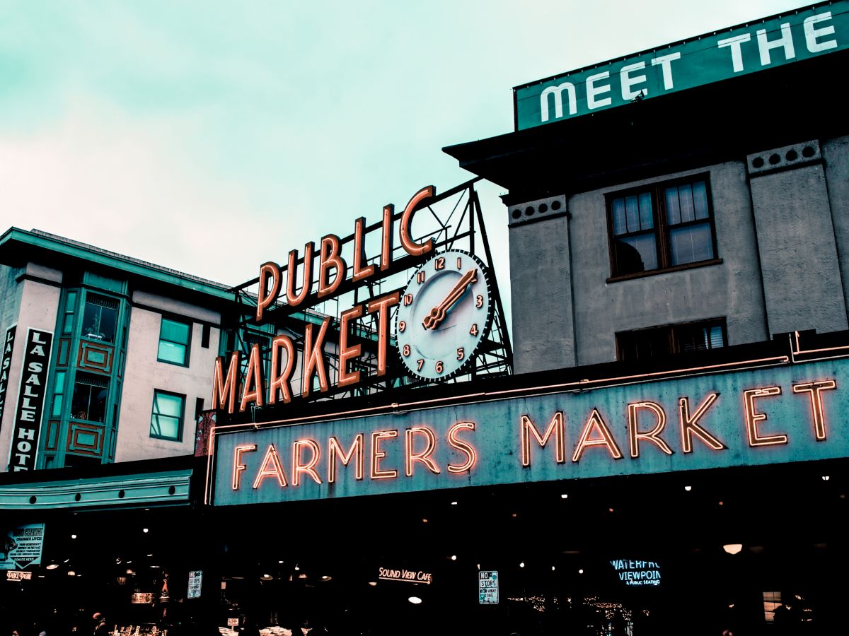 The image shows an iconic farmers market sign with a clock, and surrounding buildings under a cloudy sky.