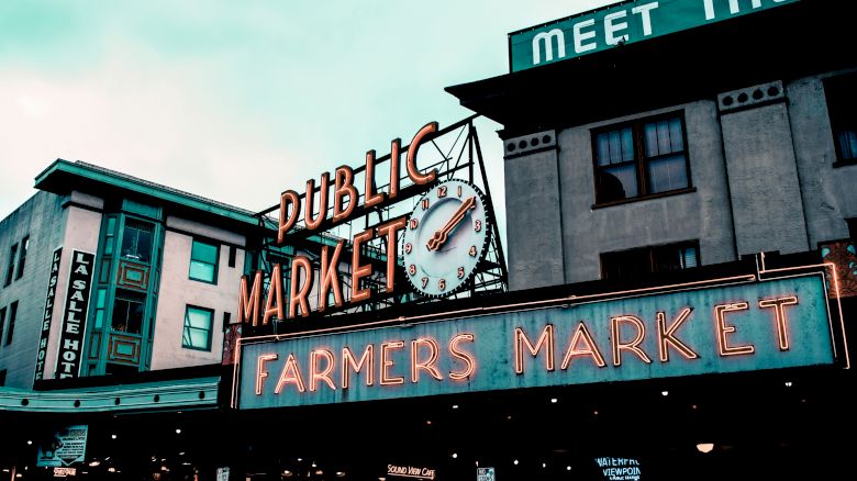 The image shows an iconic farmers market sign with a clock, and surrounding buildings under a cloudy sky.