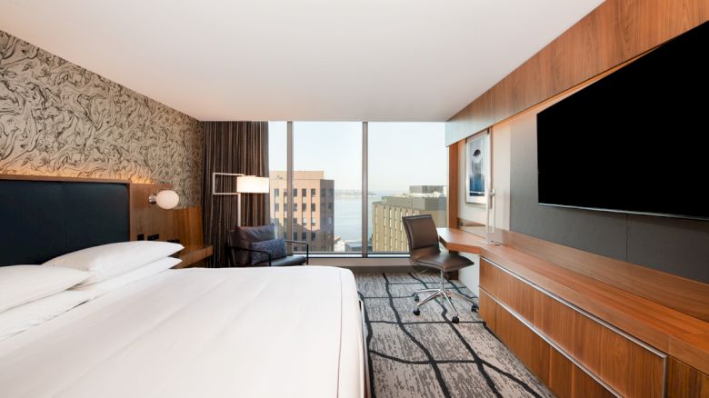 A modern hotel room with a large bed, armchair, desk, chair, and TV, featuring a city view through a window.