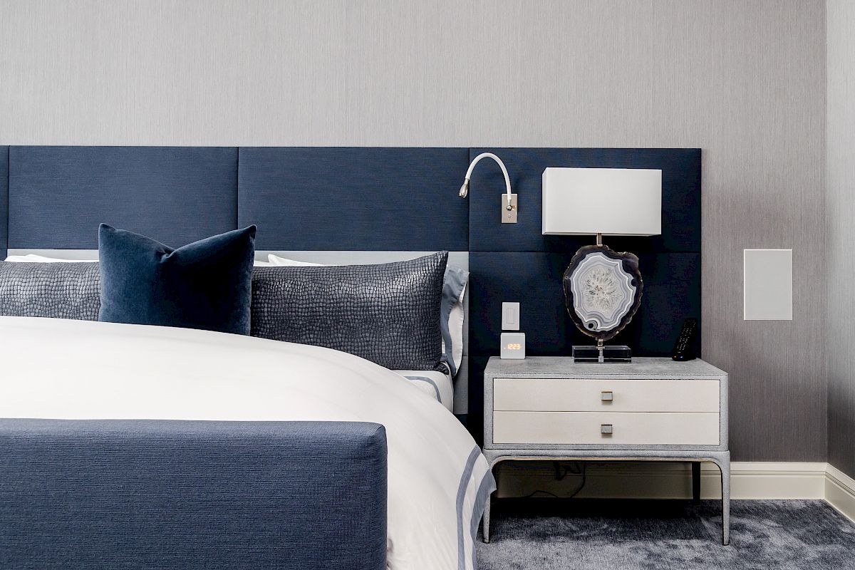 A modern bedroom with a navy upholstered bed, plush pillows, and a nightstand with a lamp and decorative items.
