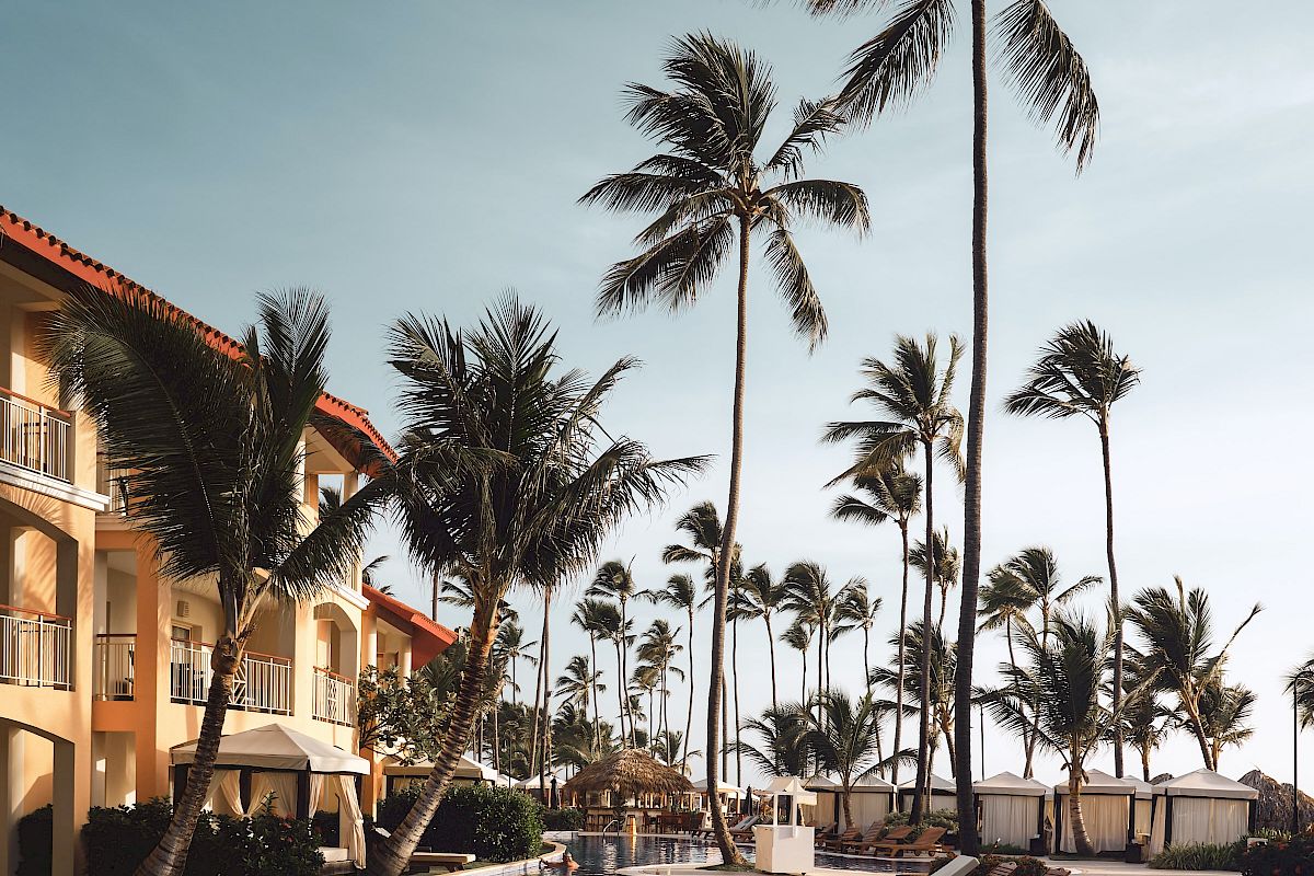 A resort poolside area with palm trees, lounge chairs, and a building in the background under a clear blue sky.