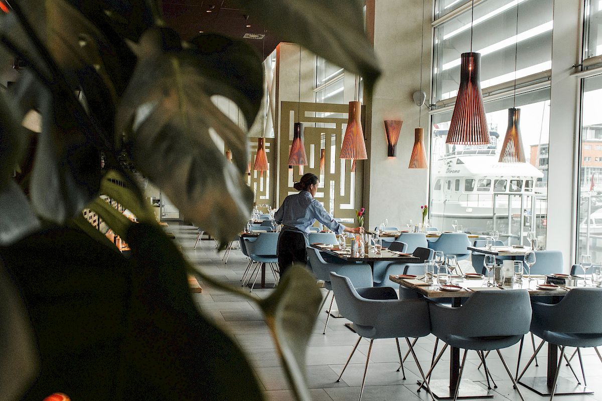 A modern restaurant with stylish decor, empty tables, and a person setting up. Large windows offer ample natural light.