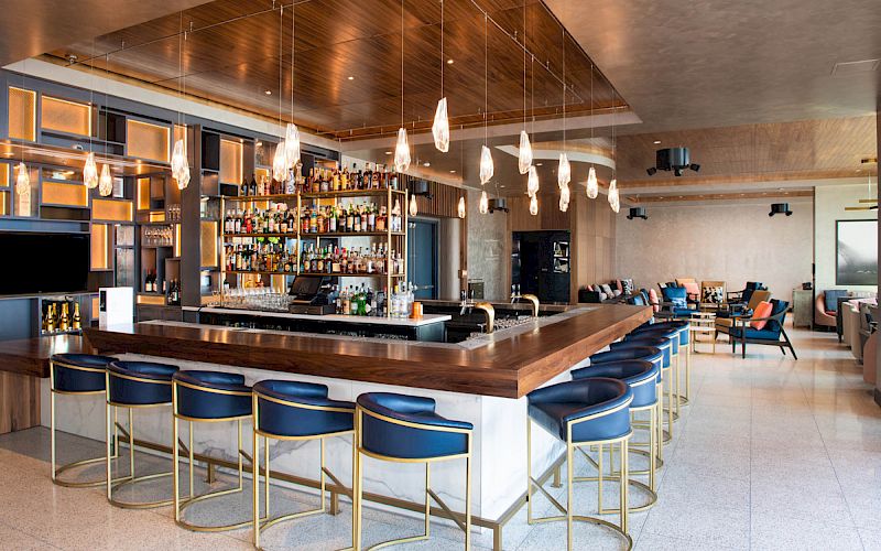 A modern bar with a sleek counter, blue stools, hanging lights, and a well-stocked liquor shelf in a spacious lounge area.