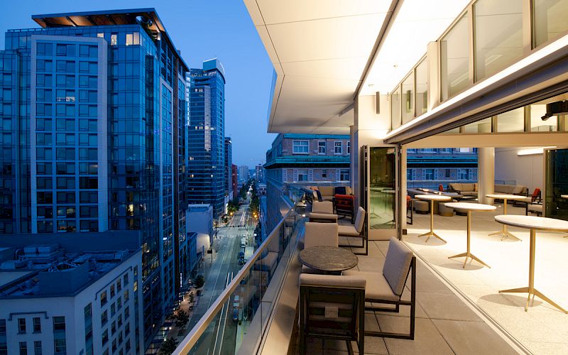 A modern rooftop terrace with tables and chairs overlooks a vibrant cityscape featuring tall buildings and a lit street below at dusk.