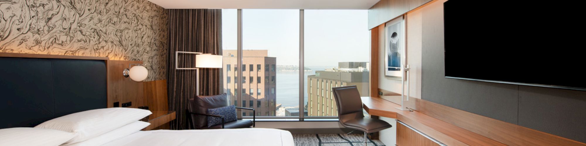 A modern hotel room with a large bed, desk, TV, and a cityscape view through the window, featuring patterned walls and contemporary decor.